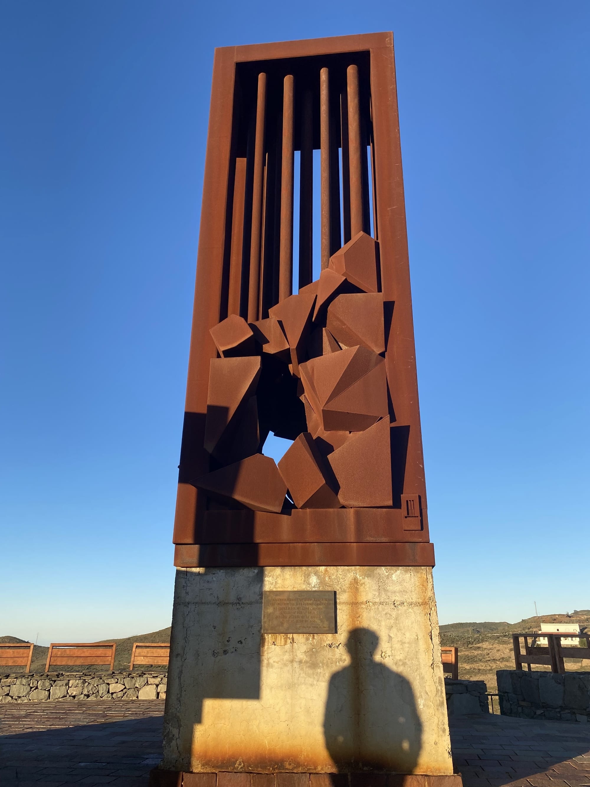 Photo of the taller of the two iron sculptures against a blue sky. A plaque is visible on its concrete base. The shadow of a man who appears to be reading it is projected onto the concrete.