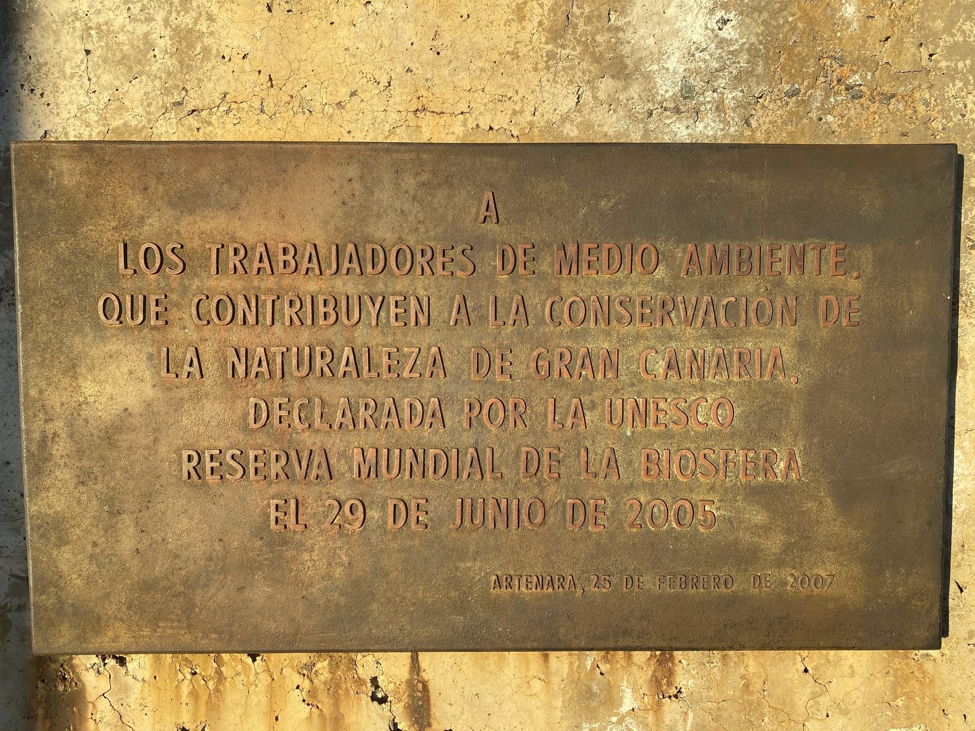 Photo of the plaque referenced throughout the article. Spanish text: “A los trabajadores del medio ambiente, que contribuyen a la conservación de la naturaleza de Gran Canaria, declarada por la UNESCO Reserva Mundial de la Biosfera el 29 de junio de 2005. Artenara, 25 de febrero de 2007.”