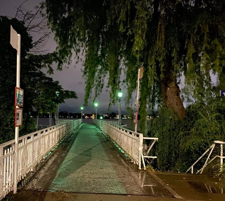 A wet asphalt path lined with a white railing stretches toward a lake beneath a midnight sky. The path glistens under three mint green streetlamps.