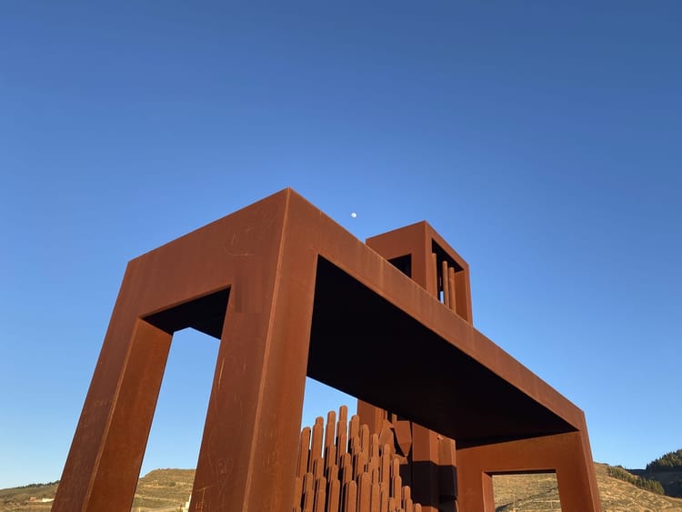 Two boxy, rust-colored metal sculptures in golden, late-afternoon light. In the background, a grassy hilltop and bright blue sky. The moon rises just above the monument.