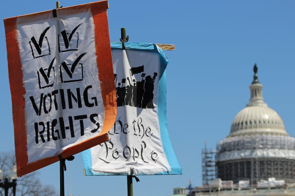 A close-up of two cloth protest signs on long poles waving against a blue sky, the dome of the U.S. Capitol in the background. One sign reads “VOTING RIGHTS,” the other “We the People.”