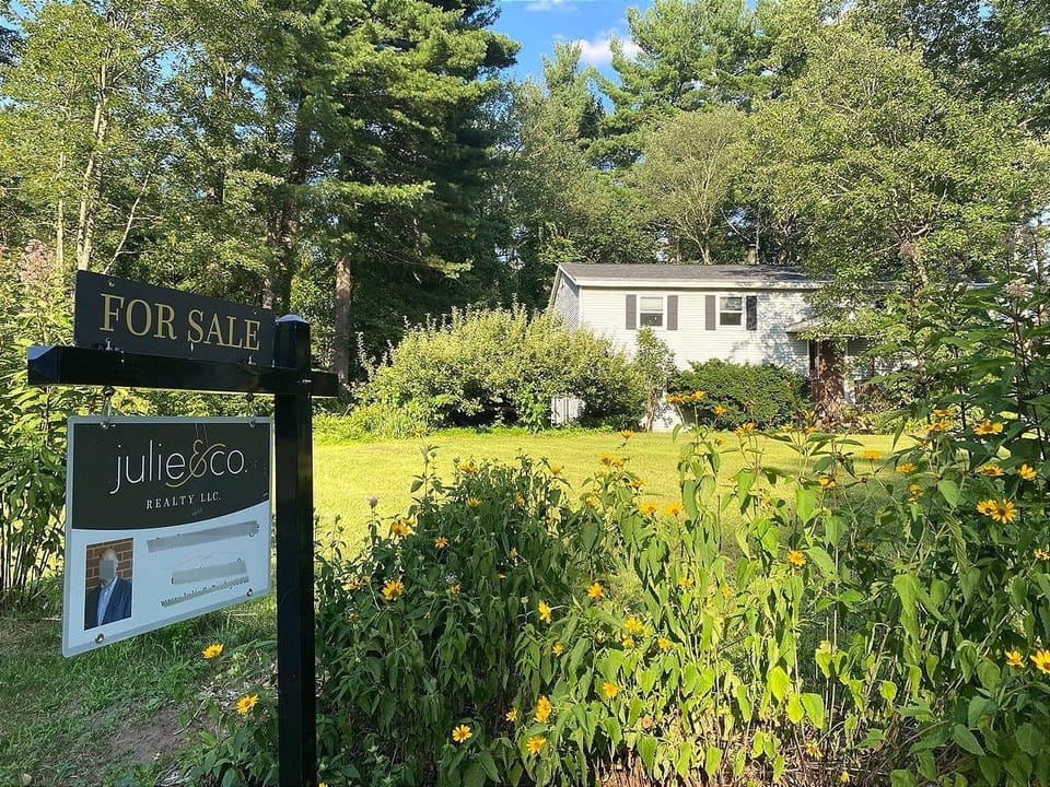 Foreground: A black-and-white “FOR SALE sign” with yellow flowers around it. Background: A green lawn and white house with black shutters on a sunny day.