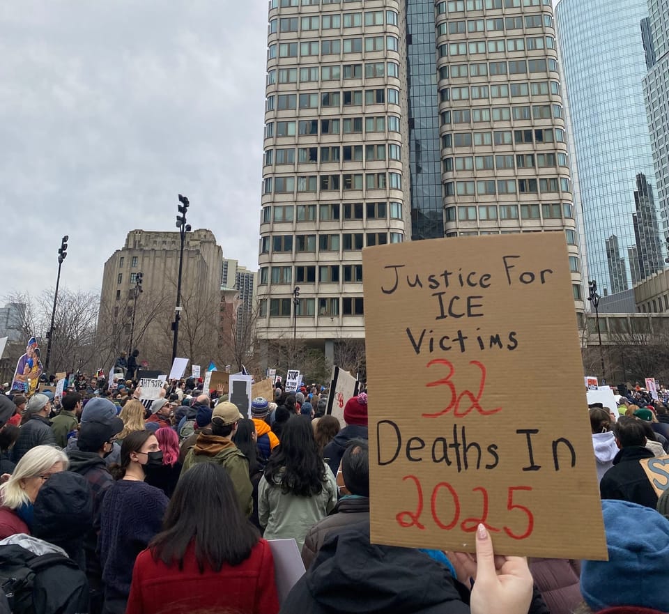 View of hundreds of people bundled against the cold at a rally in Boston. Two skyscrapers in the background. A sign in the foreground reads “Justice For ICE Victims 32 Deaths in 2025.”